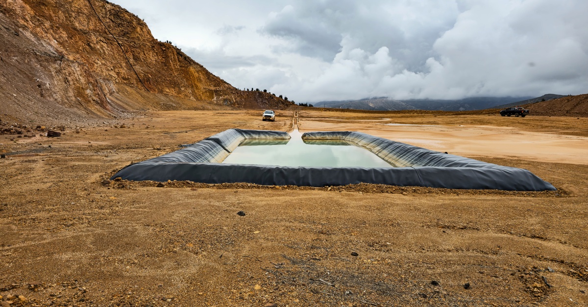 A black-lined rectangular retention pond filled with water set in a barren mining area with trucks in the distance.
