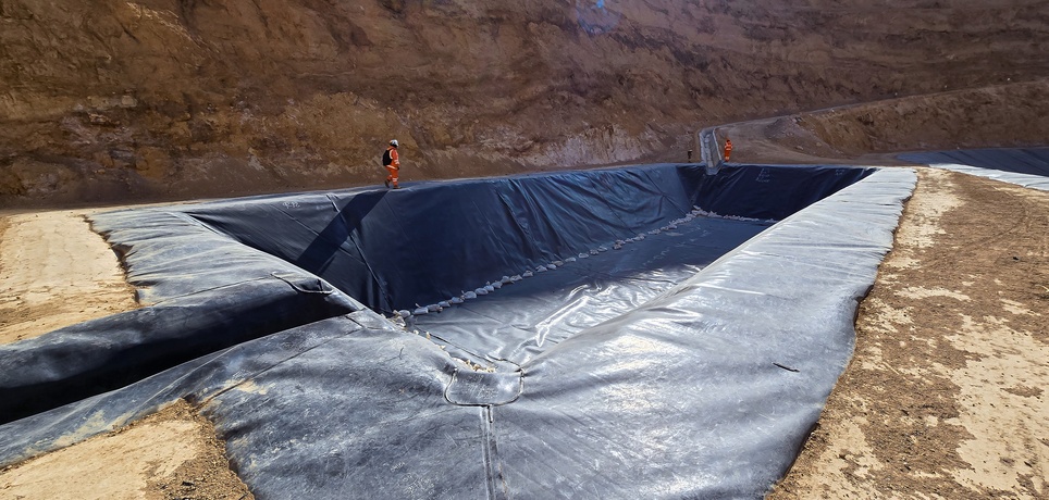 A large black geomembrane liner stretched across a barren excavation site with workers standing on the edges.