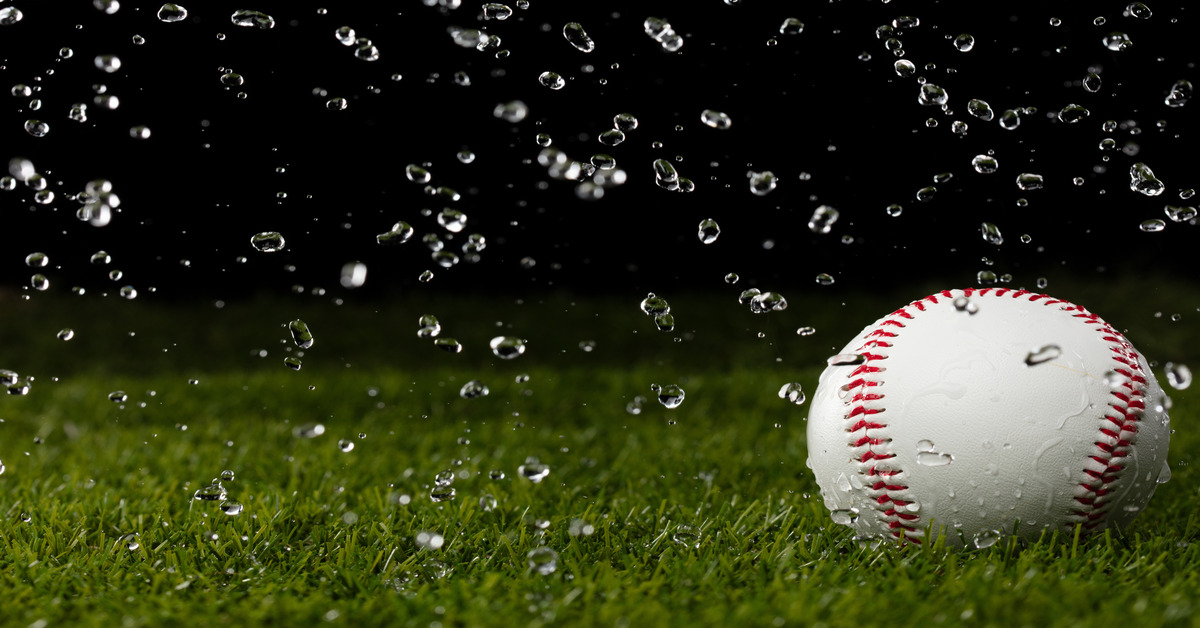 A close-up of a clean baseball sitting in the grass with rain droplets falling. The background is black.