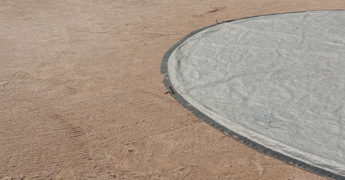 A large, circular gray tarp covering the plates of a baseball field. It is secured with anchors into the dirt.