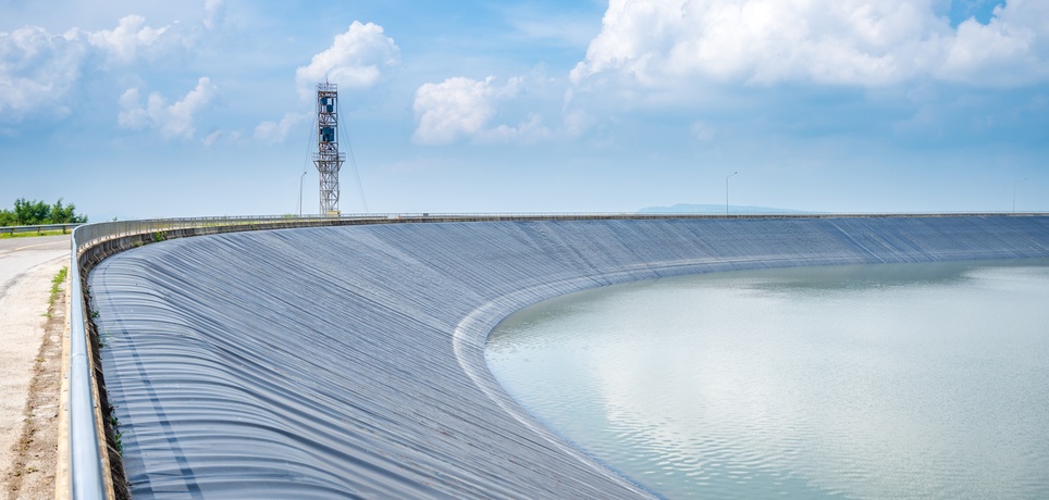 A curved black liner containing a wide lake reservoir under blue skies with a tall metal structure nearby.