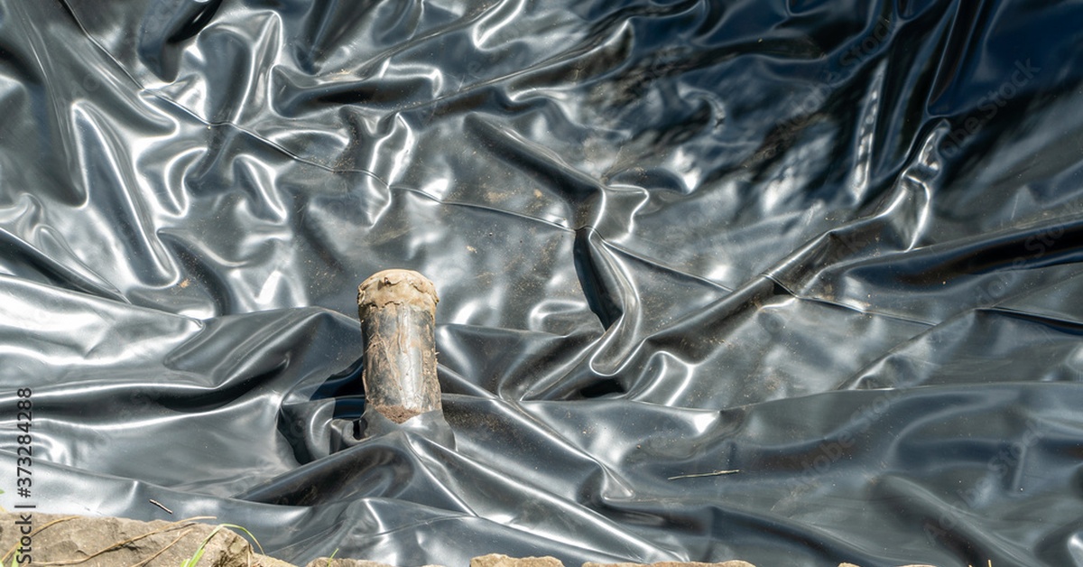 A large black pond liner laid out in an empty ditch with a pipe inserted through the liner to deliver water.