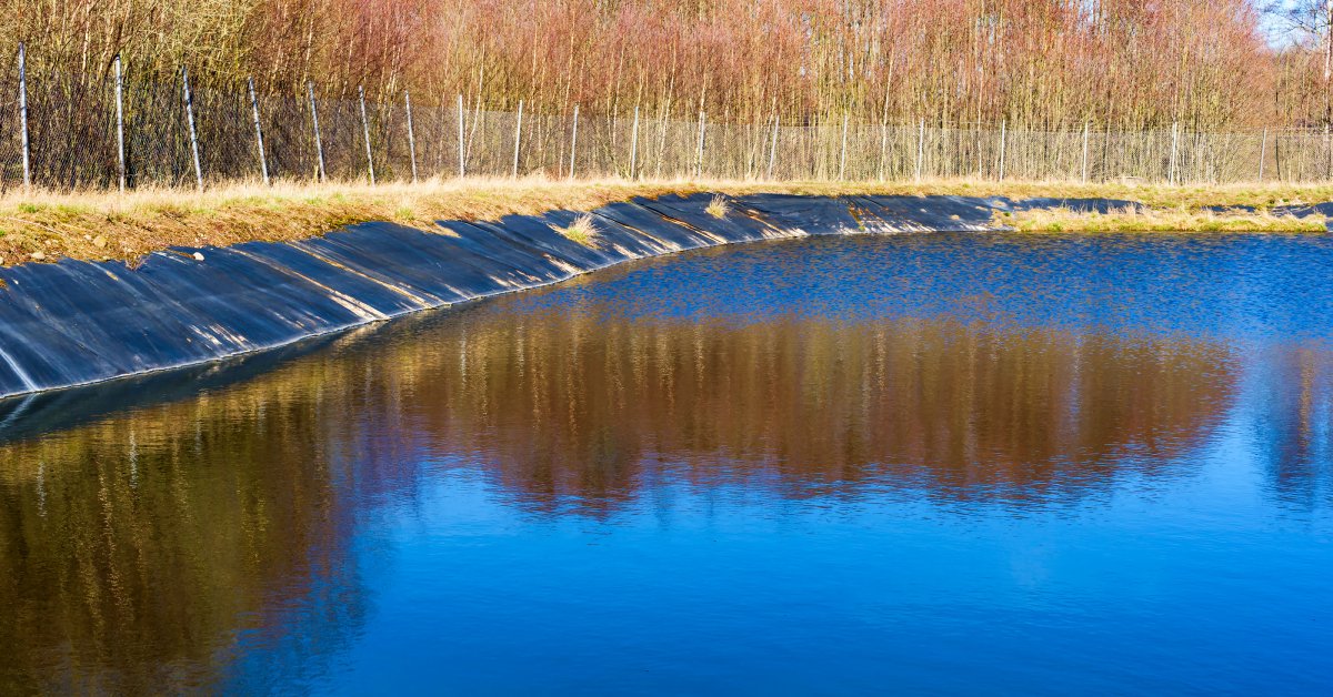 An artificial pond contains a black liner. A fence, bare trees, and brown grass surround the pond.