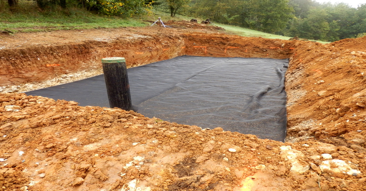 A large, rectangular hole contains a black pit liner covering the floor. The dirt is red clay with rocks.