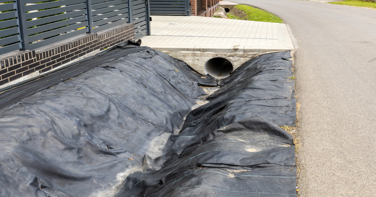 A ditch features a black liner and has dried dirt on the bottom. It has a road on one side and a black fence on the other.