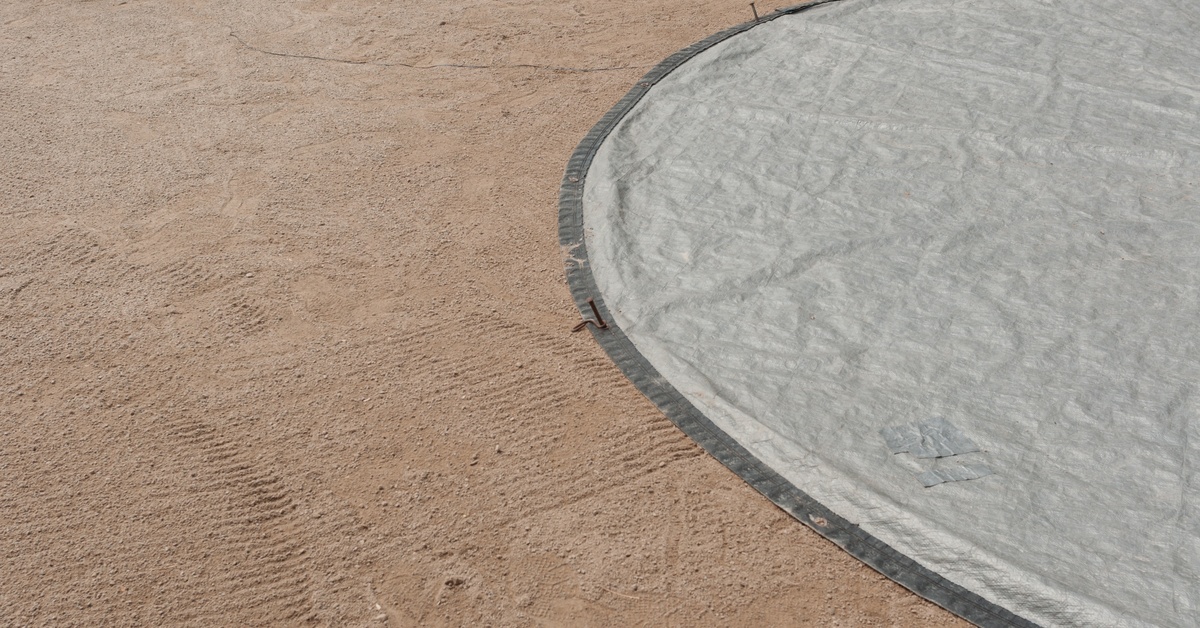 A close-up view shows a gray circular tarp covering the sandy part of a baseball field. Nails are holding down the tarp.