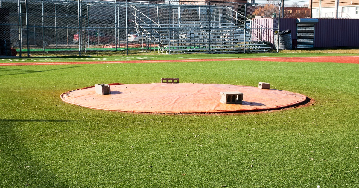 A tan-colored circular tarp covers the pitcher's mound on a baseball field. Cinderblocks weigh down the tarp.