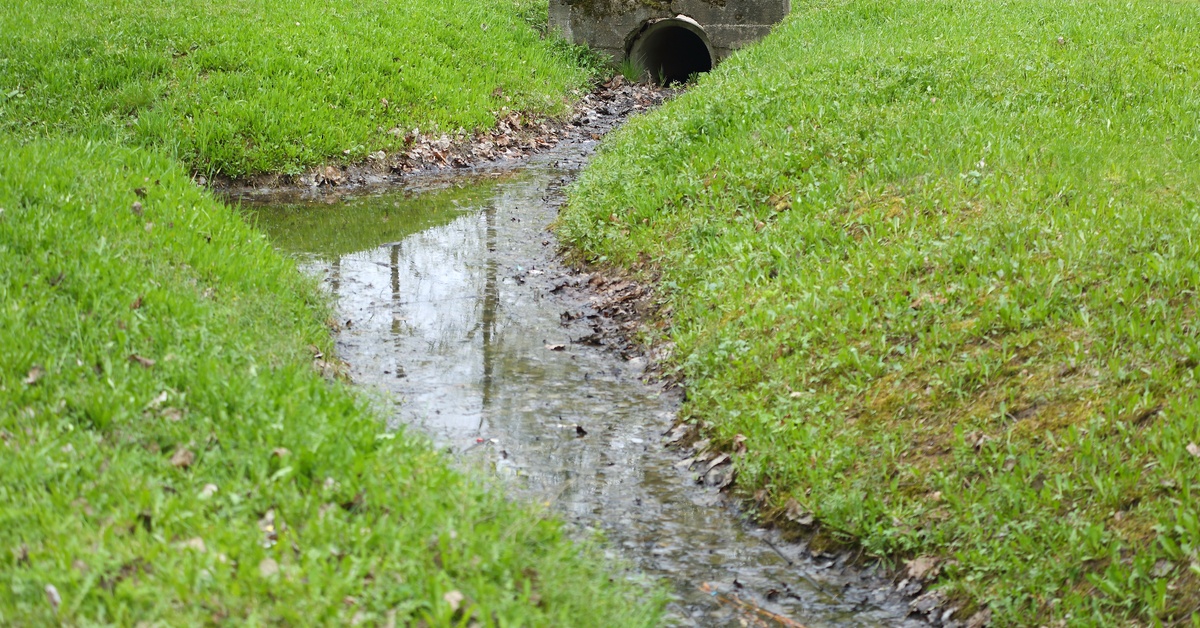 Water is flowing through a ditch that sits at the bottom of a grassy hill. A pipe helps the water flow through a small bridge.