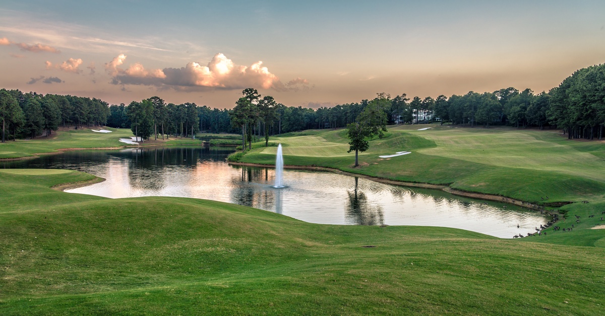 A large pond sits on a beautiful golf course. The pond has a water feature that shoots the water into the air.