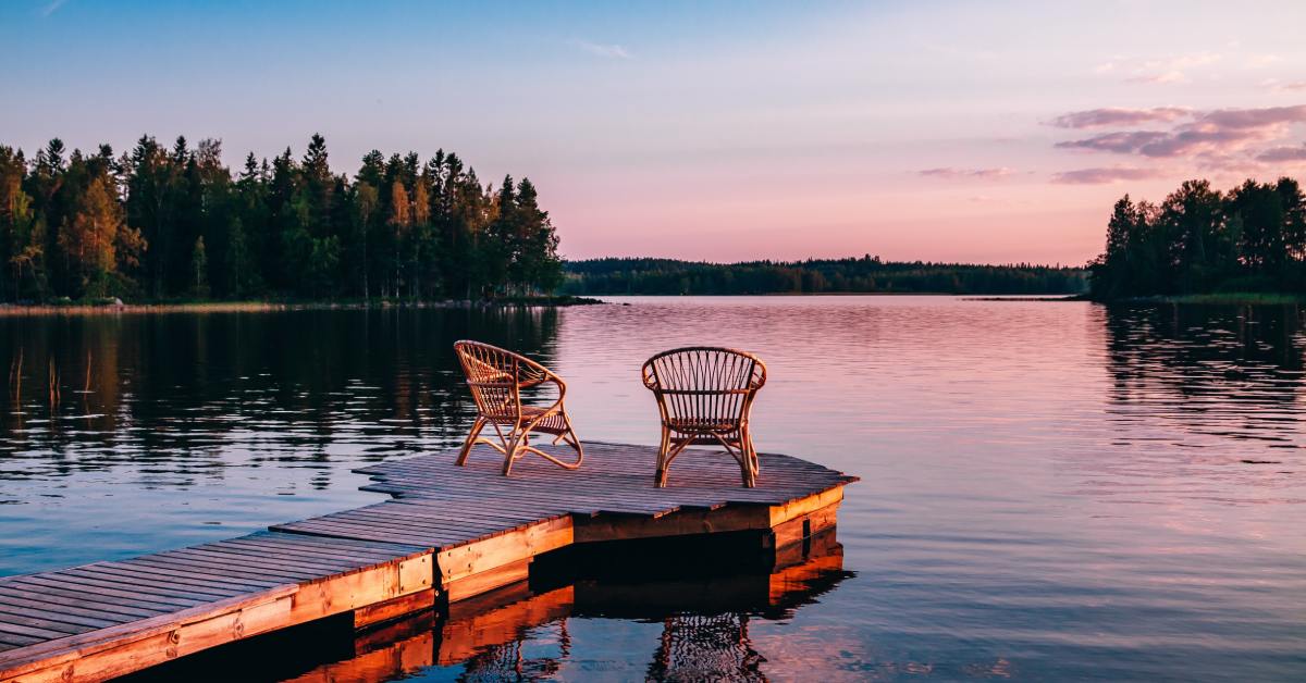 wo wooden chairs sit at the end of a wooden dock overlooking a lake. The sunset is making the sky blue, purple, and pink.