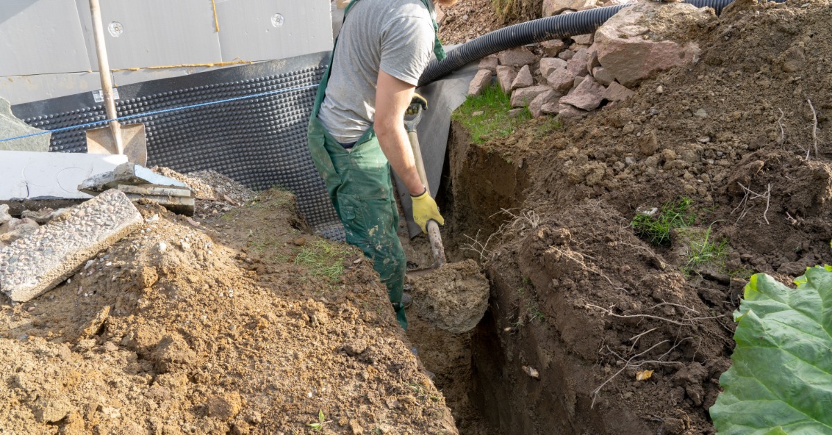 A man wearing a gray T-shirt and green overalls using a shovel to remove dirt from the ditch he's digging.