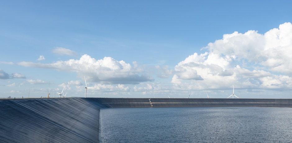 An artificial lake features a black lake liner and is full of water. Several white wind turbines are in the background.