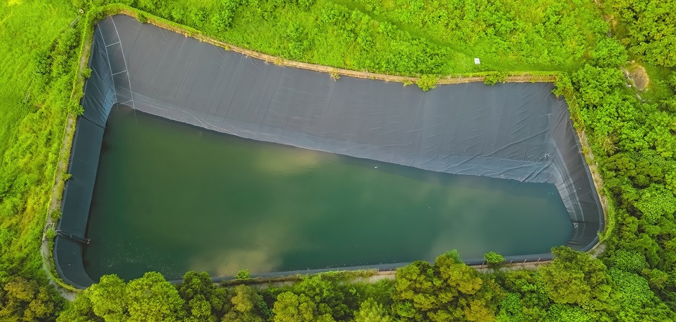 An aerial view shows a large pond with a black liner that's surrounded by trees. The pond contains water on the bottom.