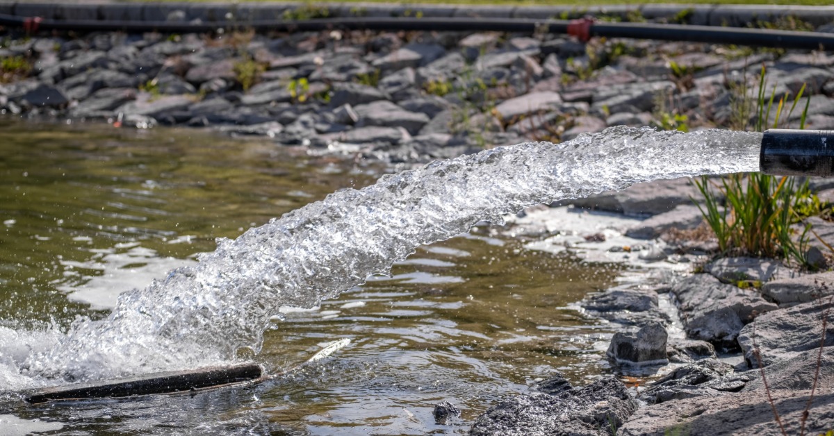 A close-up view shows water flowing quickly out of a pipe and into a pond. Rocks are at the bottom of the pond.