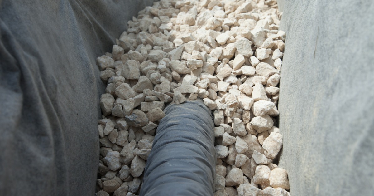 A drainage ditch features a liner that goes up both sides and has white rocks on the bottom. A black pipe goes into the rocks.
