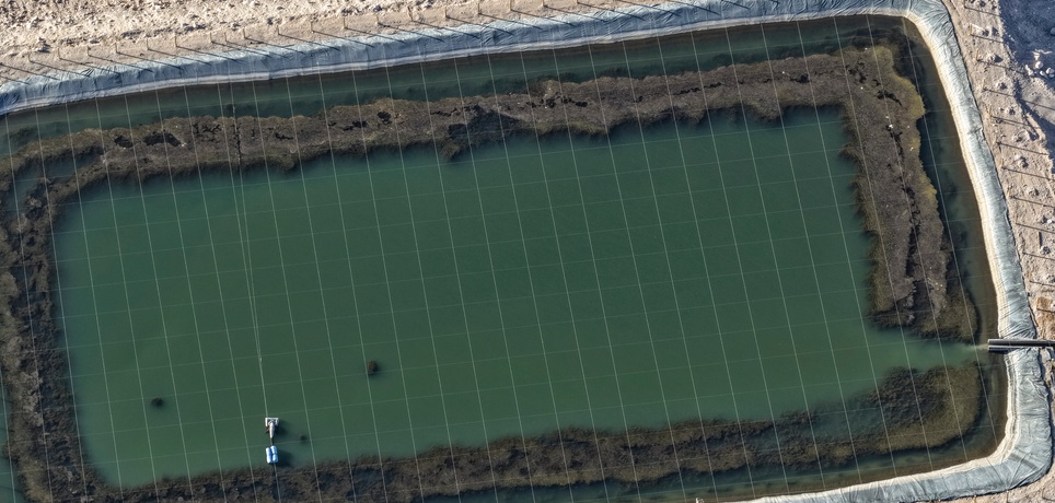 An aerial view shows a large pond filled with water. The pond has a black liner and is surrounded by dirt and sand.