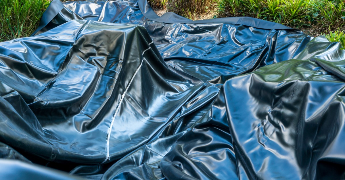 A close-up view shows a black pond liner that sits crinkled on grassy area. The feet of people is in the background.