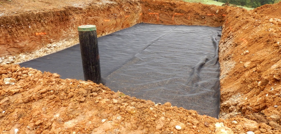 A large pit is dug in a red clay soil, and a black pit liner lines the bottom of the pit. A black pipe emerges from the side.