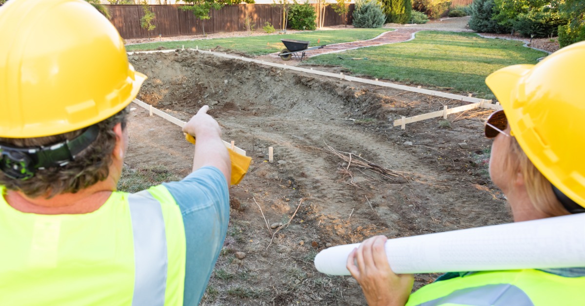 A man and a woman wearing neon-yellow safety vests and yellow hard hats observe a large hole in the ground.