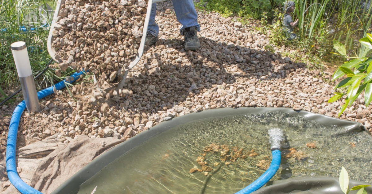 A person is using a wheelbarrow to dump a bunch of rocks at the edge of a pond that's filled with water.