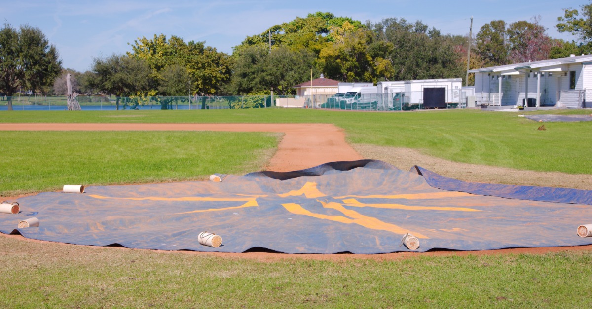 A blue tarp is covering a batter's box and the surrounding sand on a baseball field. The tarp has sand on it.