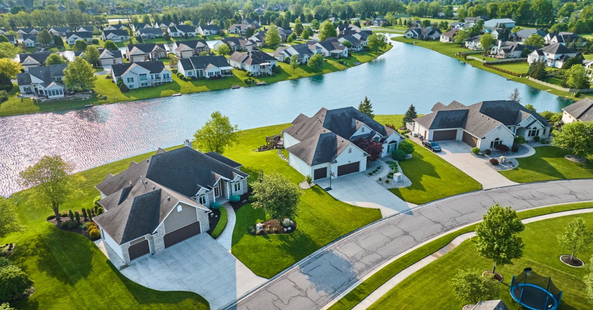 An aerial view shows a pond surrounded by the backyards of newer homes. The grass in the yards is green.