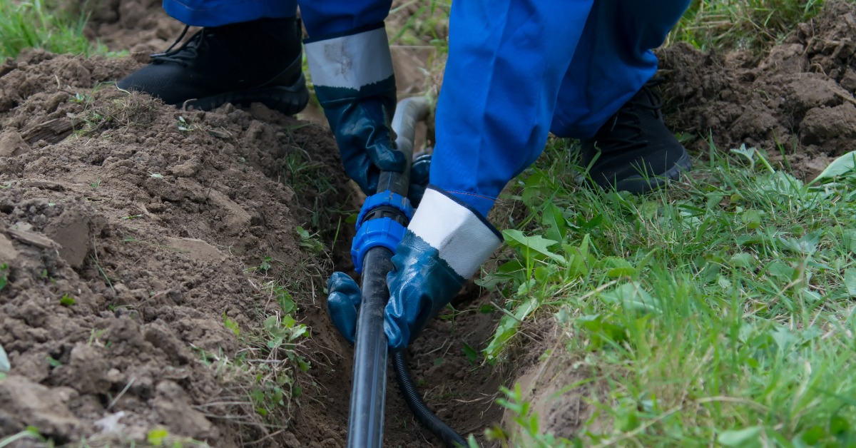 A person wearing a blue work suit and work gloves is placing a black plastic pipe in a dirt ditch.